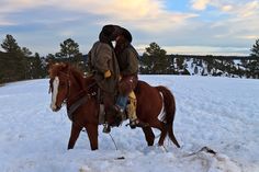 two people riding on the back of a brown horse in the middle of a snow covered field