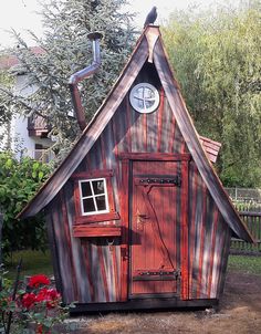 a small wooden building with a clock on the top and side window, in front of some trees