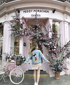 a woman standing in front of a pink building with lots of flowers on the outside