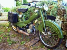 an old green motorcycle parked next to a stone wall and shrubbery in the background