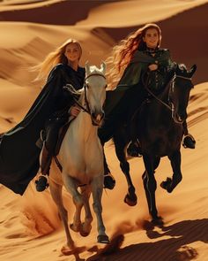 two women are riding horses in the sand dunes, one is wearing a black cape