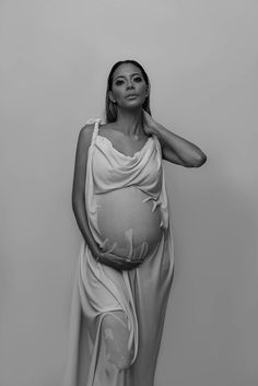 a black and white photo of a pregnant woman in a draped dress with her hands on her head