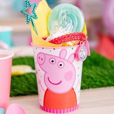 a peppa pig cup filled with candy and other party supplies on a table next to pink cups