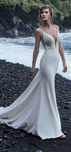 a woman in a white wedding dress standing on the rocks by the water with her hands behind her back