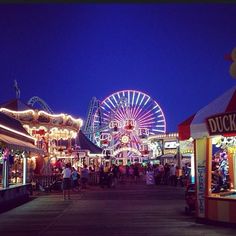 the carnival is lit up at night and people are walking on the boardwalk near ferris wheel