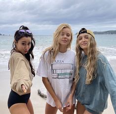 three girls standing on the beach with their hands in each other's pockets and looking at the camera