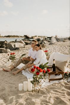 a man and woman sitting on top of a sandy beach next to the ocean with candles in front of them