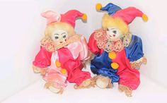 two stuffed clowns sitting next to each other on a white surface with one wearing a pink and blue outfit