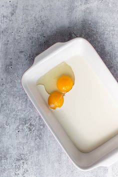 two eggs in a white square dish on a gray table top with one egg broken