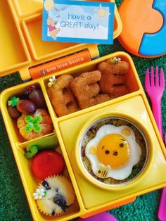 a yellow lunch box filled with food on top of a green floor next to plastic utensils