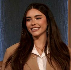 a woman with long brown hair wearing a tan suit and gold hoop earrings smiles at the camera