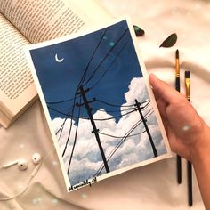 a hand holding an open book with power lines and moon in the sky behind it