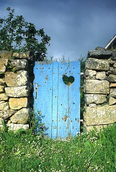 an old stone wall with a blue door and heart cutout in the middle, surrounded by green grass