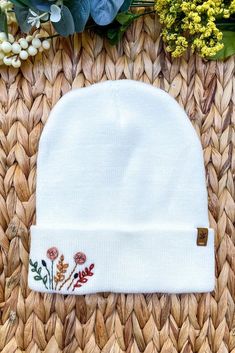 a white beanie with flowers on it sitting next to some green leaves and flowers