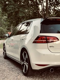 the back end of a white car parked in front of some trees and gravel road