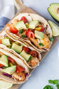 three fish tacos with avocado, tomatoes and cilantro on a cutting board