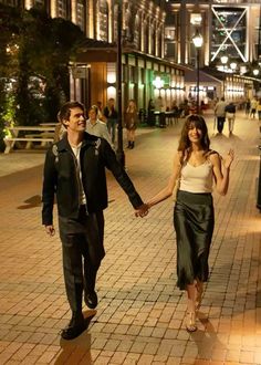 a man and woman holding hands walking down the street in front of buildings at night