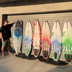 a man standing next to five surfboards in front of a garage door with his hand on the board