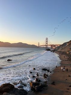 people are walking on the beach next to the water and suspension bridge in the background