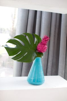 a pink flower in a blue vase on a white shelf next to a curtained window