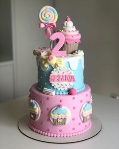 a pink and blue birthday cake with two cupcakes on it's top