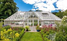 a greenhouse with lots of plants and flowers in it's front yard, surrounded by greenery