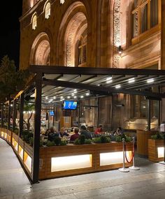 people are sitting at tables in front of a building that has lights on the outside