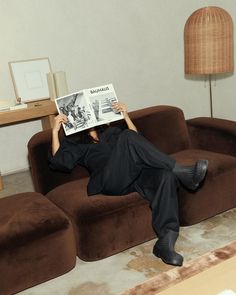 a man laying on top of a brown couch next to a table with a lamp