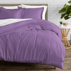 a bed with purple comforter and pillows in front of a potted green plant