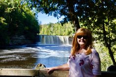 a woman standing next to a wooden fence near a river and forest with waterfall in the background