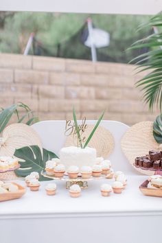 a table topped with lots of cupcakes next to a palm leaf covered wall