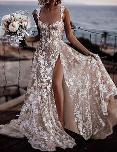 a woman in a white dress standing on a wooden deck next to the ocean and holding a bouquet