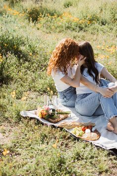 two women sitting on a blanket in the middle of a field with food and drinks