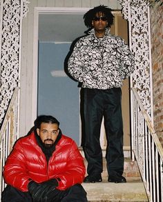 two young men sitting on the steps in front of a house, one wearing a red jacket