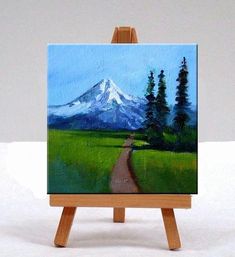 an easel with a painting of a road leading to a snowy mountain in the distance