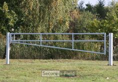 an empty metal fence in the middle of a field with trees and bushes behind it