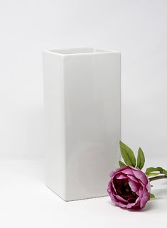 a white square vase with a pink flower in front of it on a white background