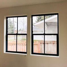 two windows with black frames in an empty room next to a wooden fence and white house