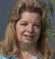 a woman with blonde hair and blue shirt smiling at the camera while standing in front of a tree