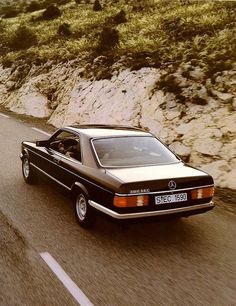 an old mercedes benz coupe driving down the road in front of a mountain side cliff