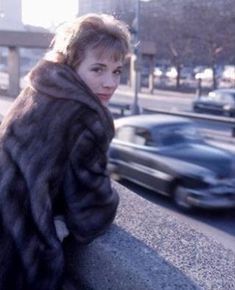 a woman in a fur coat leaning against a wall on the side of a road