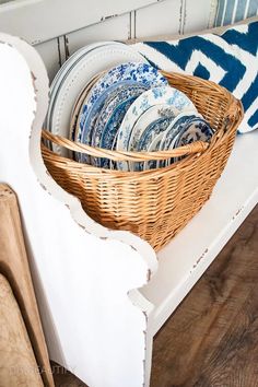 a basket filled with plates sitting on top of a white bench next to a blue and white pillow