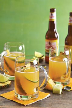 two glasses filled with yellow liquid next to bottles and slices of lime on a wooden table