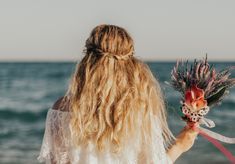 a woman standing on the beach with her hair blowing in the wind and holding flowers