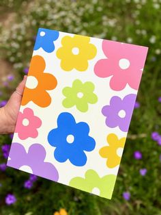 a hand holding up a colorful flowered card in front of purple and yellow flowers