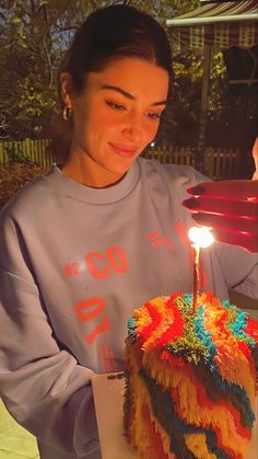 a woman holding a birthday cake with a lit candle on it and wearing a gray sweatshirt