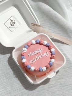 a pink birthday cake with white and blue frosting in a box next to a fork