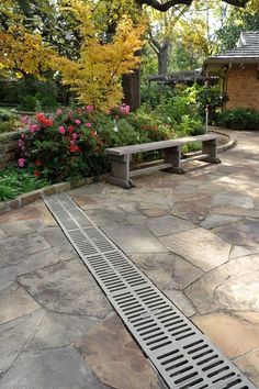 a stone walkway in front of a house with flowers and trees on the other side