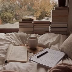 an open book, cup and pen on a bed in front of a window with trees outside