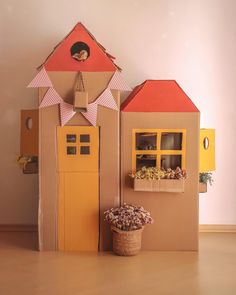 two cardboard houses with flowers in front of them and a potted plant on the floor